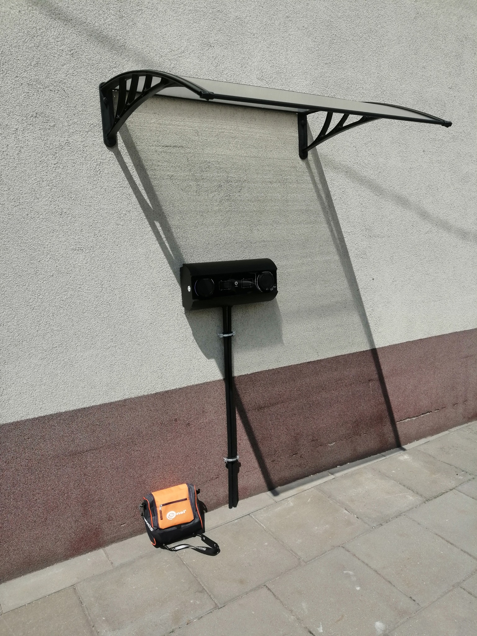 Czarny głośnik zewnętrzny z kamerą, zamontowany na elewacji pod zadaszeniem, obok torba narzędziowa Sonel na chodniku.