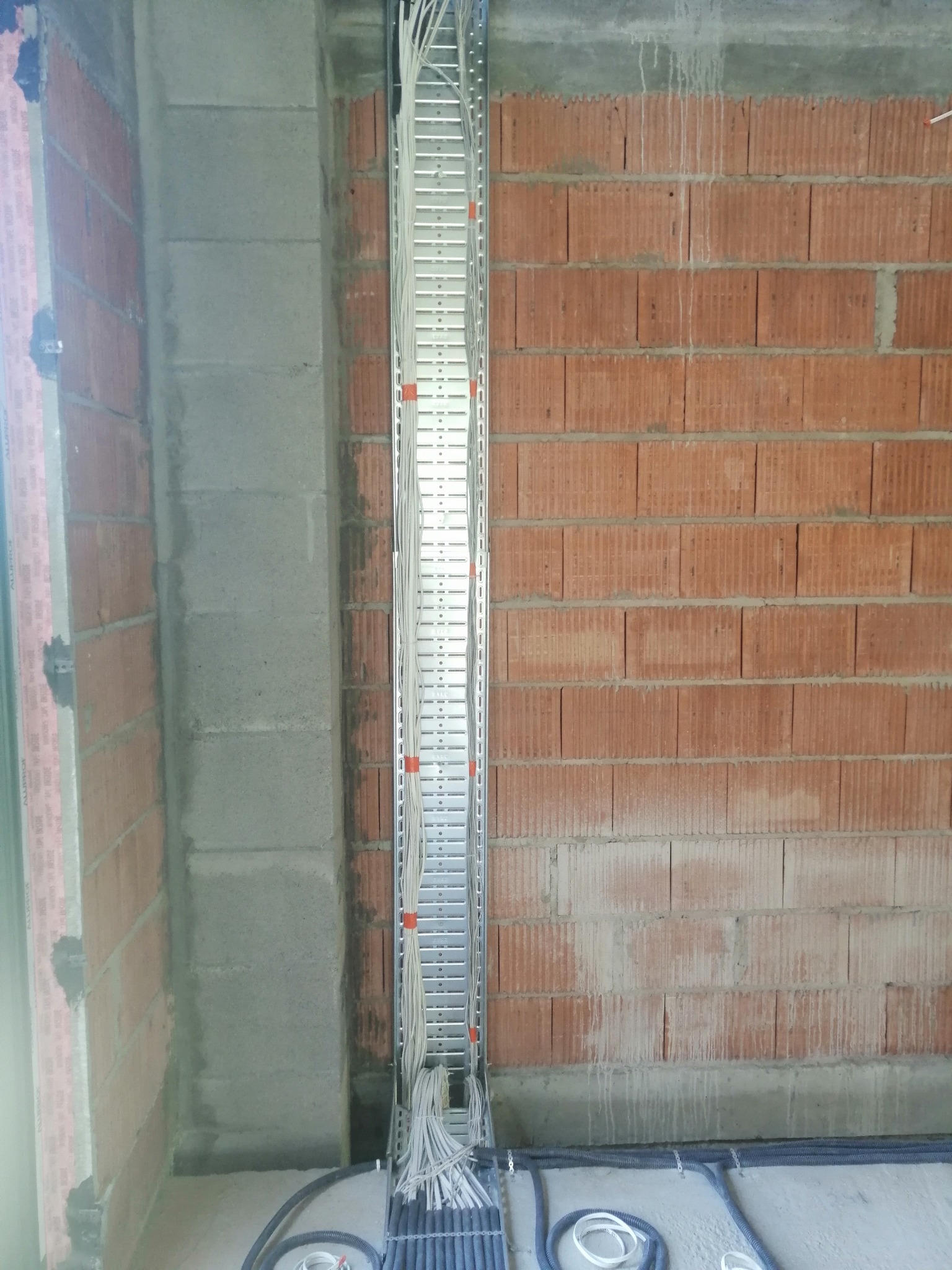 Wewnętrzna instalacja elektryczna w trakcie montażu: koryto kablowe pionowo zamocowane do surowej ściany z cegły, z widocznymi przewodami i peszlami na podłodze.