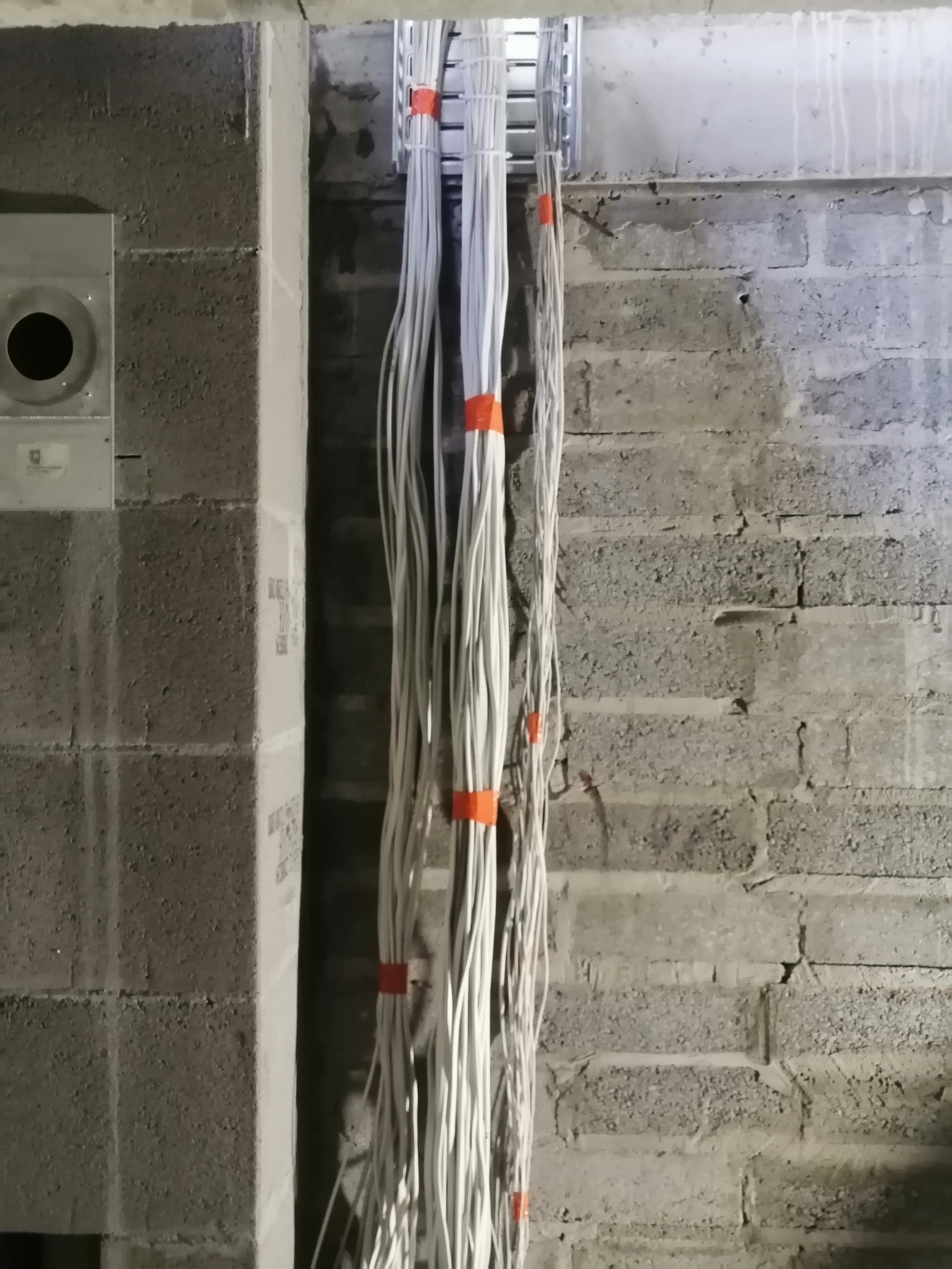Pionowy układ białych przewodów elektrycznych, zabezpieczonych pomarańczowymi opaskami, wychodzących z metalowej koryta kablowego, na tle surowej ściany z betonowych bloczków.