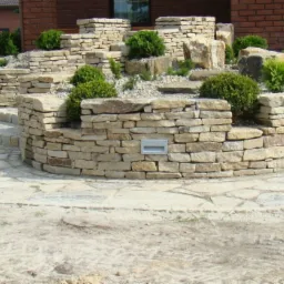 Kamienna kaskada ogrodowa z niskimi murkami, schodami i nasadzeniami kulistych krzewów, oświetlona punktowymi lampami wbudowanymi w murki.