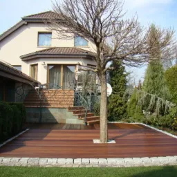 Drewniany taras z wkomponowanym drzewem, obrzeża z jasnego kamienia, schody z balustradą z motywem roślinnym prowadzące do domu.