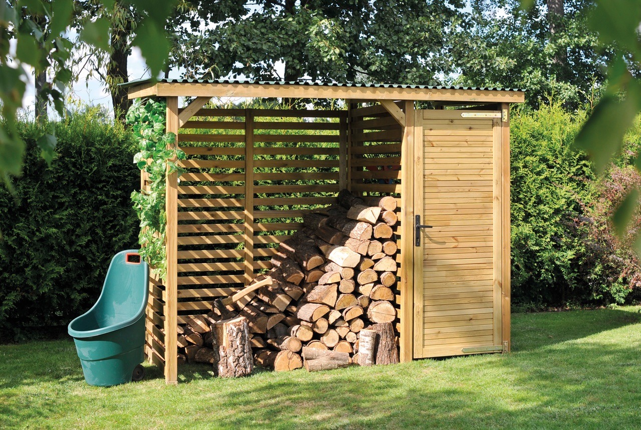 Drewniana konstrukcja z zadaszeniem, służąca jako skład drewna, z ułożonymi polanami, zielonym wózkiem ogrodowym i pnączem na słupie, na tle trawnika i zieleni.