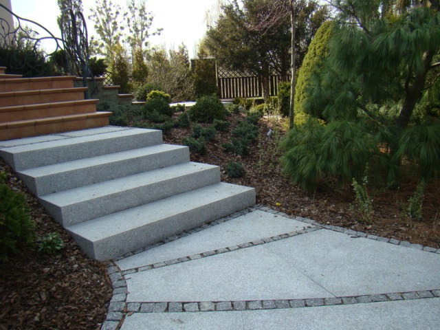 Granitowe schody ogrodowe prowadzące do domu, z dekoracyjnym obramowaniem z kostki brukowej i roślinnością w tle.