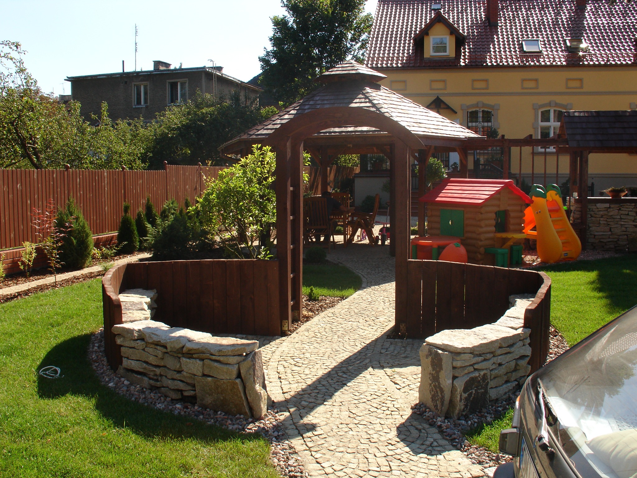 Ogród z drewnianą altaną z meblami ogrodowymi, kamienną ścieżką i elementami drewnianej architektury ogrodowej, w tle dom z czerwoną dachówką oraz plac zabaw dla dzieci.