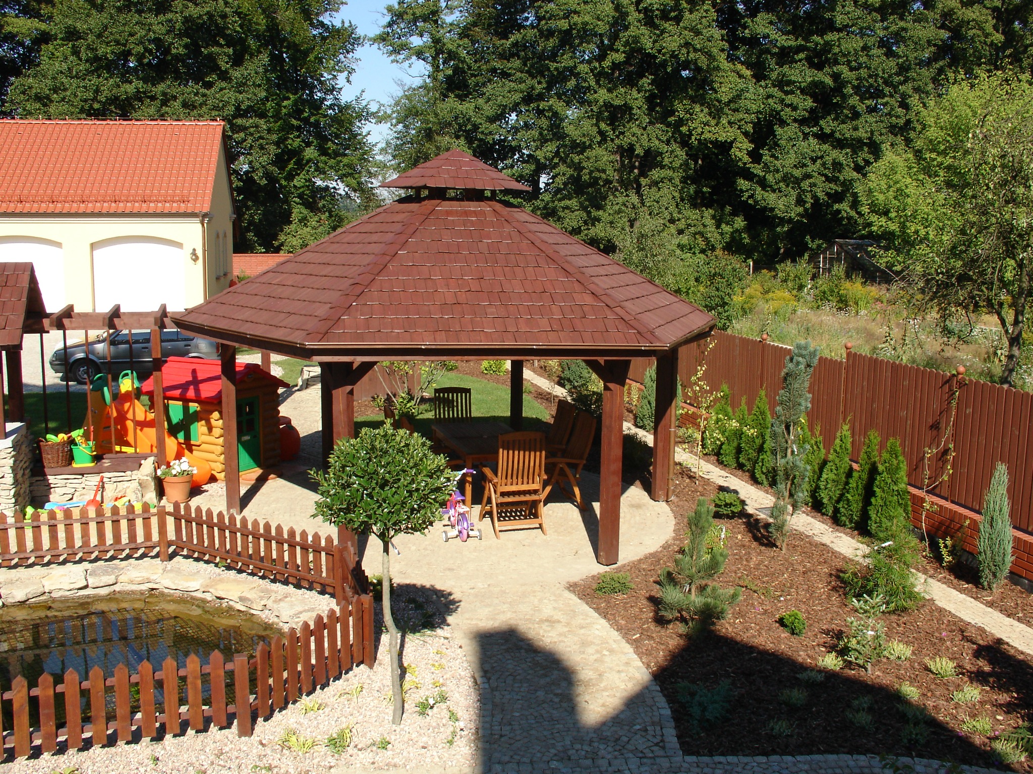 Widok z góry na brązową altanę z drewnianymi meblami ogrodowymi, małym trójkołowcem i drzewkiem w donicy, otoczoną zadbanym ogrodem z oczkiem wodnym i ścieżkami wyłożonymi kostką.