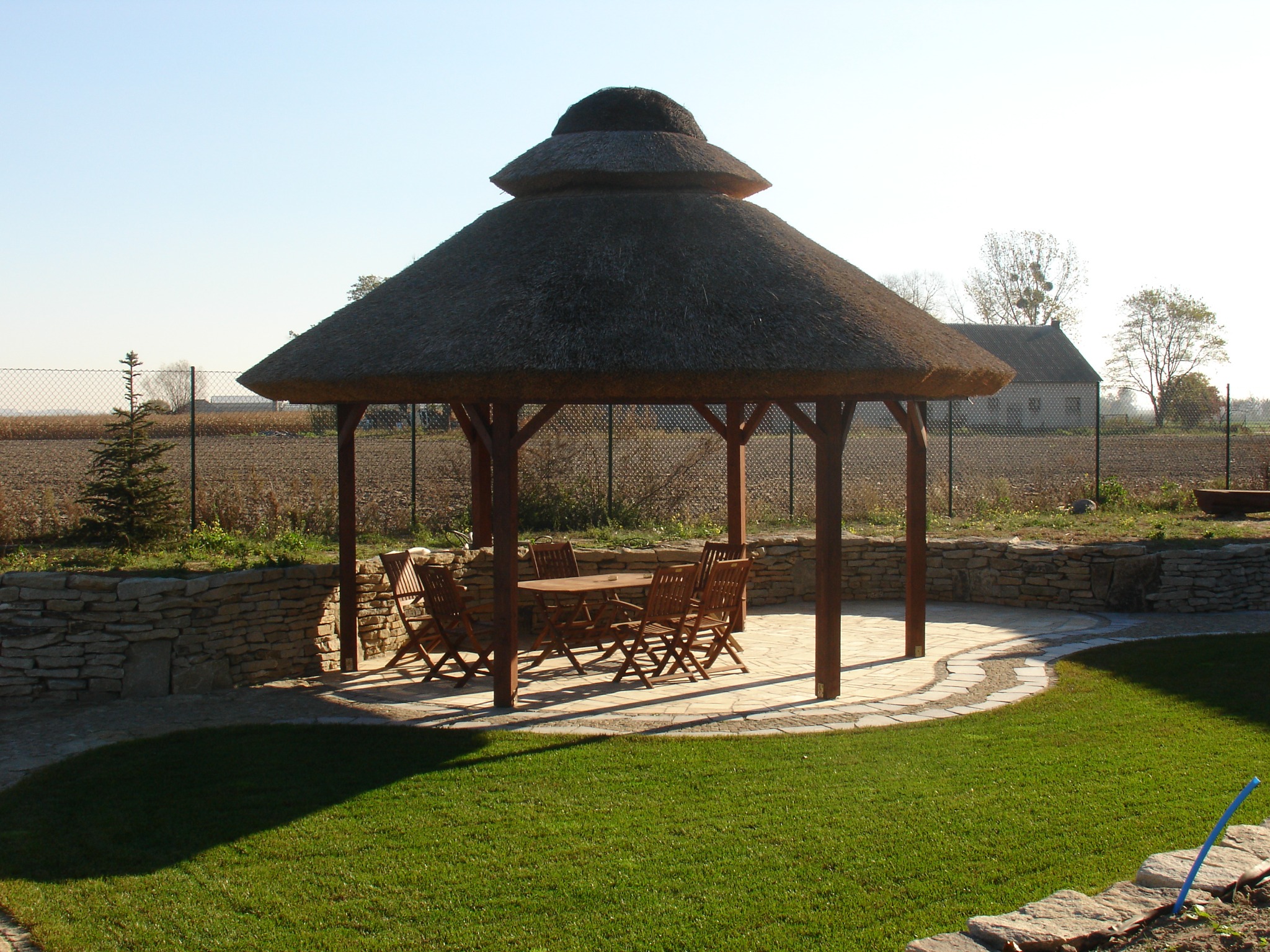 Altana z dwupoziomowym, strzechowym dachem, drewnianymi słupami i meblami ogrodowymi, otoczona kamiennym murkiem i trawnikiem na tle pola i budynku gospodarczego.