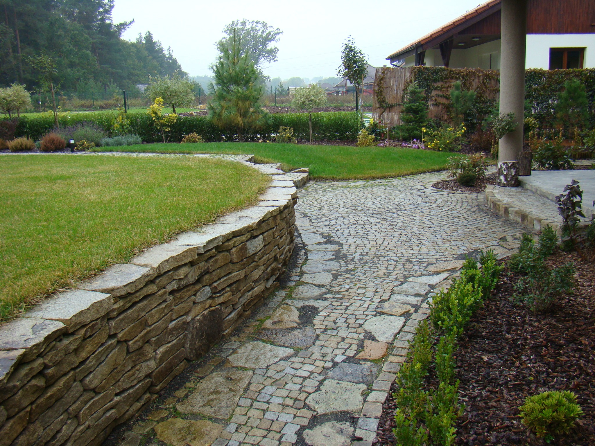 Kamienna ścieżka z kostki brukowej wijąca się przez zadbany ogród z niską kamienną ścianką, trawnikiem i różnorodną roślinnością, prowadząca w kierunku domu.