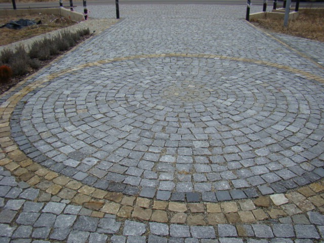 Ułożona z kostki granitowej ścieżka z dekoracyjnym okręgiem z jaśniejszej kostki, prowadząca w stronę chodnika z przydrożną roślinnością.