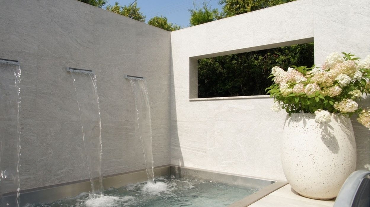 Nowoczesna aranżacja ogrodu z minimalistyczną fontanną kaskadową wbudowaną w szarą ścianę, z widokiem na zieleń przez prostokątne okno i ozdobną donicą z hortensjami.