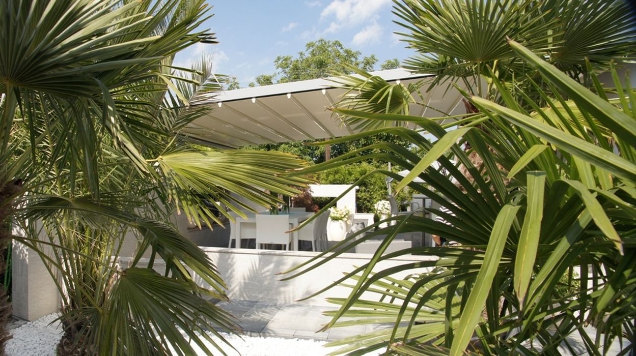 Biała pergola z rozsuwanym dachem, widoczna przez liście palmy, z białymi meblami ogrodowymi i dekoracyjnymi białymi kamykami.