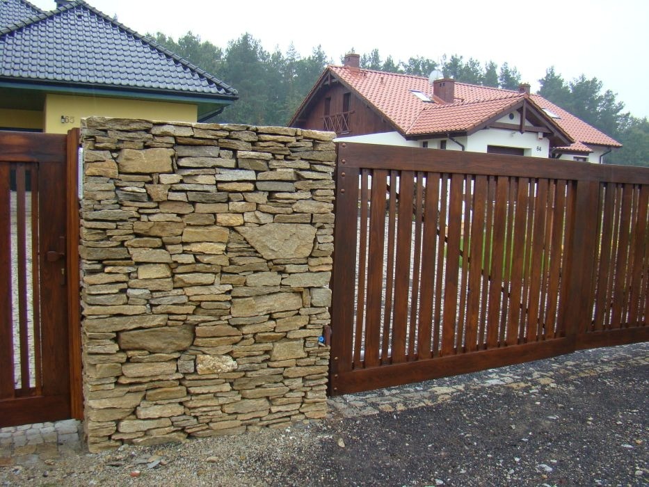 Drewniana brama wjazdowa z pionowymi szczeblami, przylegająca do kamiennego murku z nieregularnych, płaskich kamieni, na tle domów i drzew.