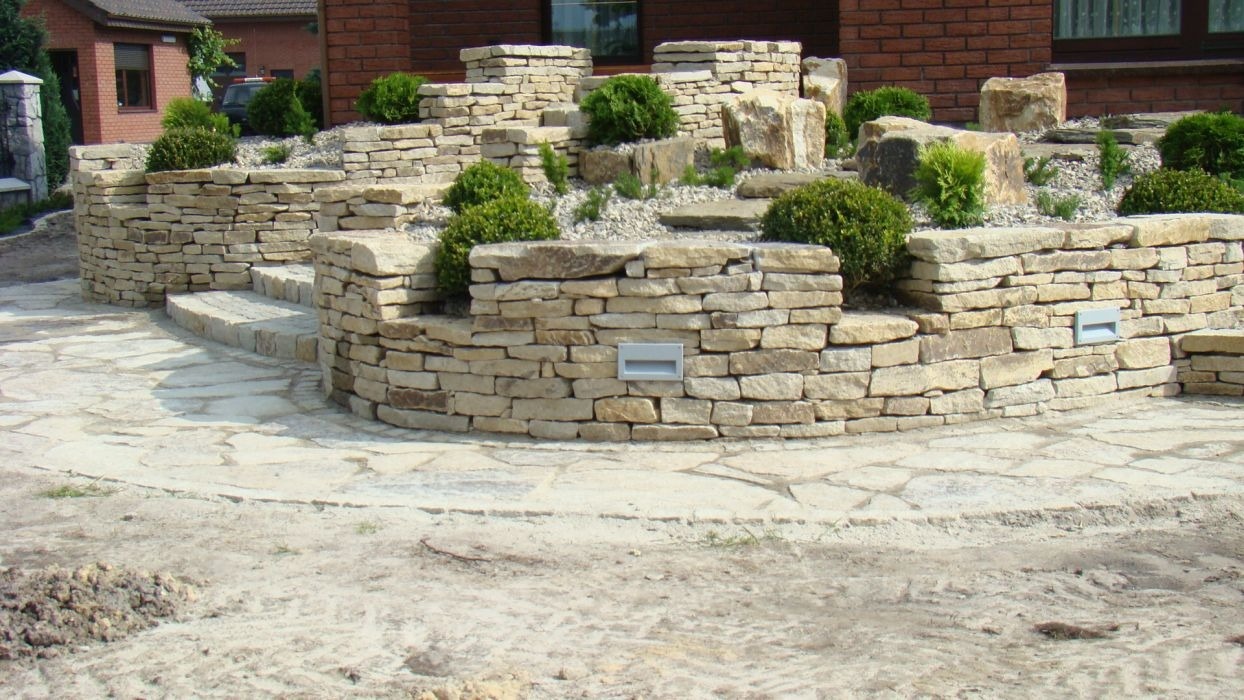 Kamienna kaskada ogrodowa z niskimi murkami, schodami i nasadzeniami kulistych krzewów, oświetlona punktowymi lampami wbudowanymi w murki.