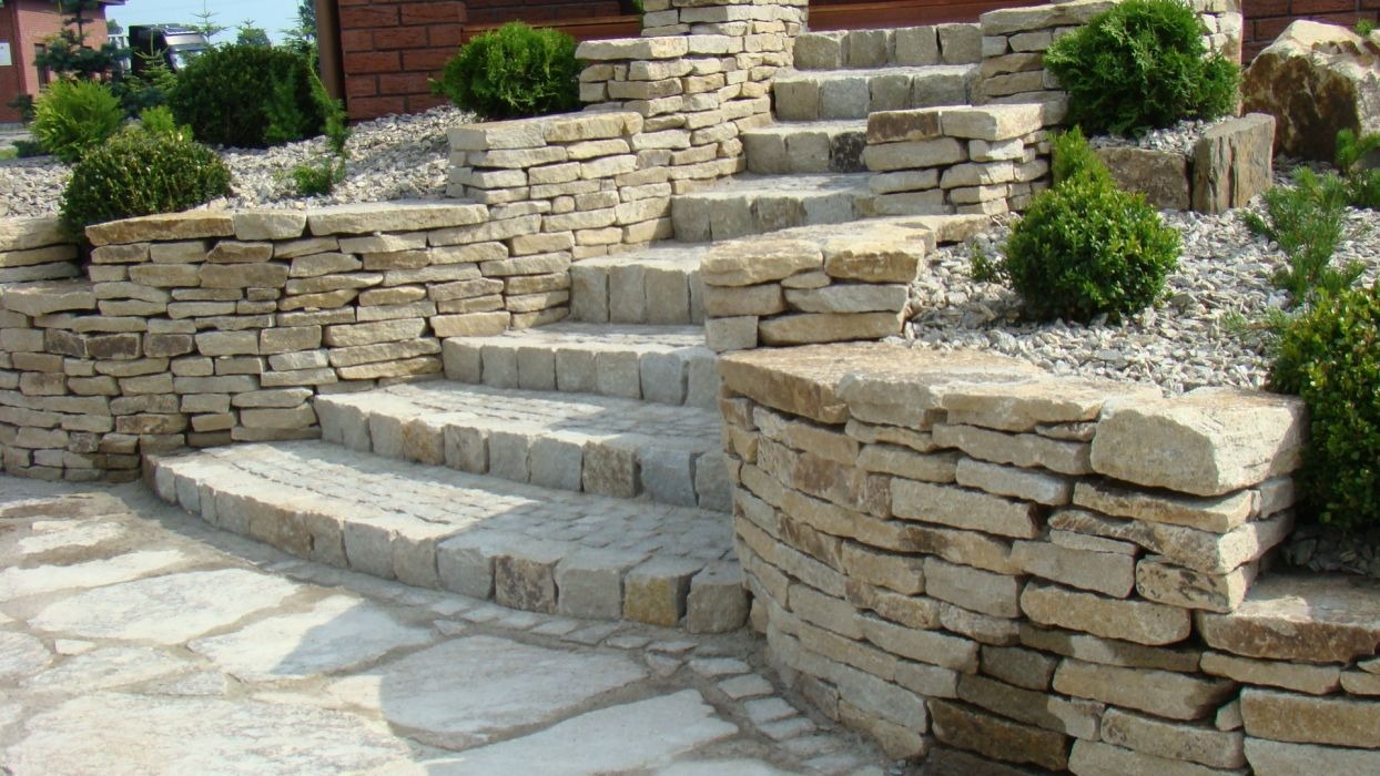 Kamienne schody ogrodowe z niskimi murkami oporowymi, obsadzone kulistymi krzewami i wysypane drobnym żwirem, prowadzące do domu z ceglaną elewacją.