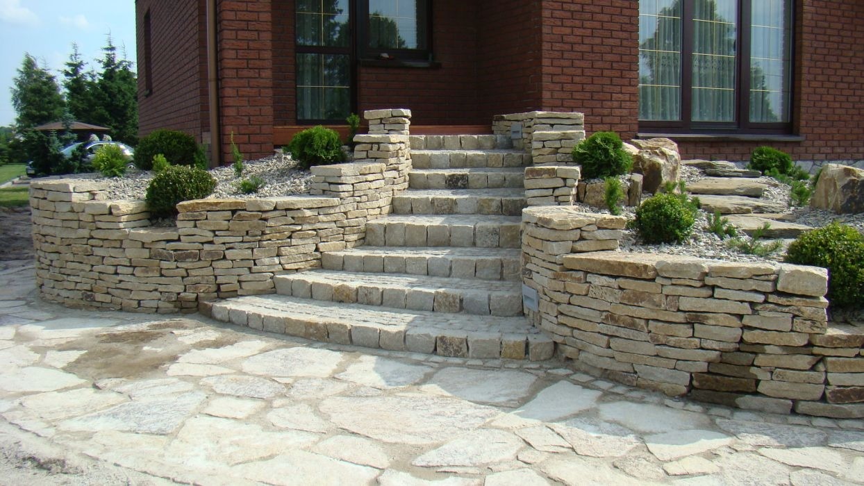 Kamienne schody wejściowe do domu z obudową z kamienia łupanego i rabatami z roślinnością ozdobną.