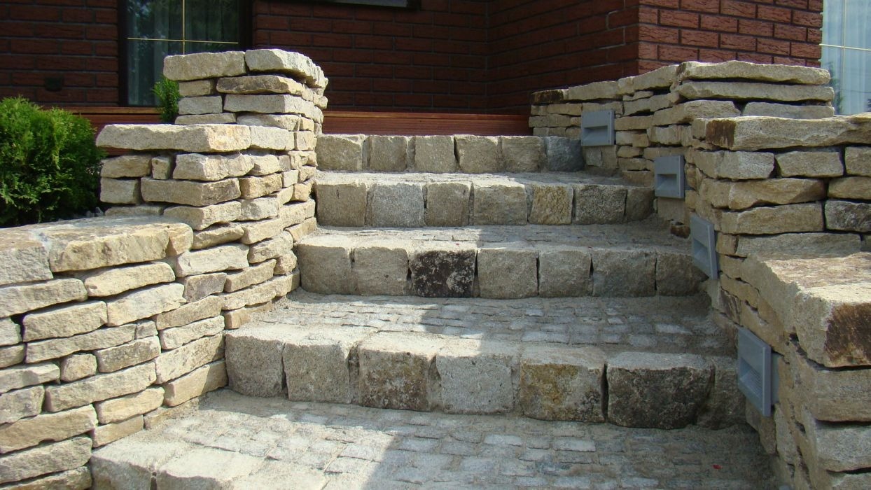 Kamienne schody zewnętrzne z podświetleniem w murze, prowadzące do domu z ceglaną elewacją i zielonym krzewem obok.