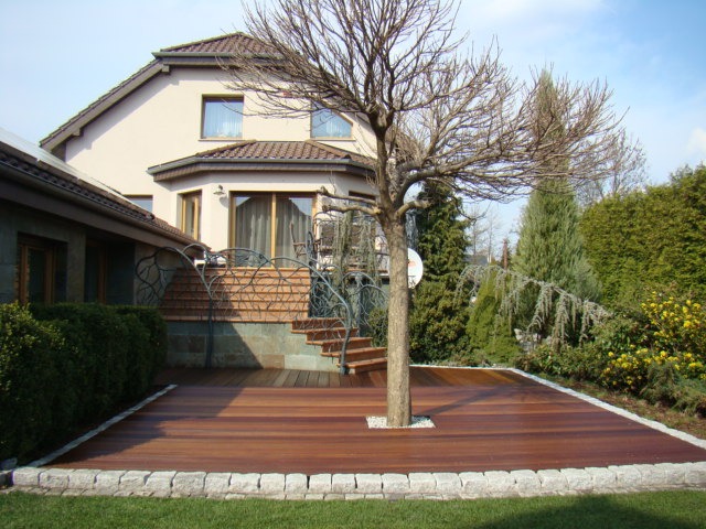 Drewniany taras z wkomponowanym drzewem, obrzeża z jasnego kamienia, schody z balustradą z motywem roślinnym prowadzące do domu.
