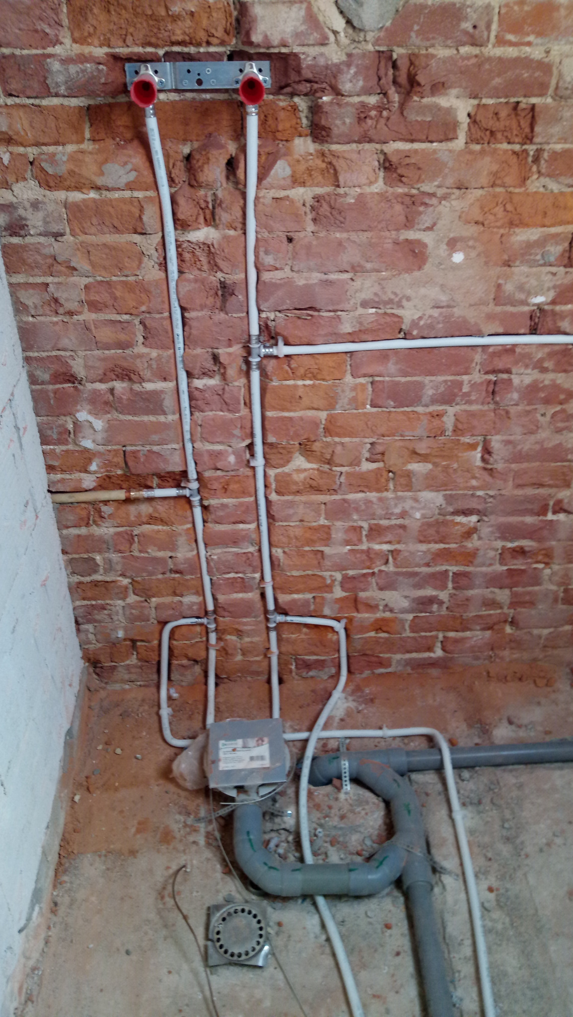 Instalacja wodna z białych rur PCV i szarych rur kanalizacyjnych na tle ściany z czerwonej cegły, widoczne przyłącza z czerwonymi zaślepkami, skrzynka elektryczna i metalowa kratka ściekowa...