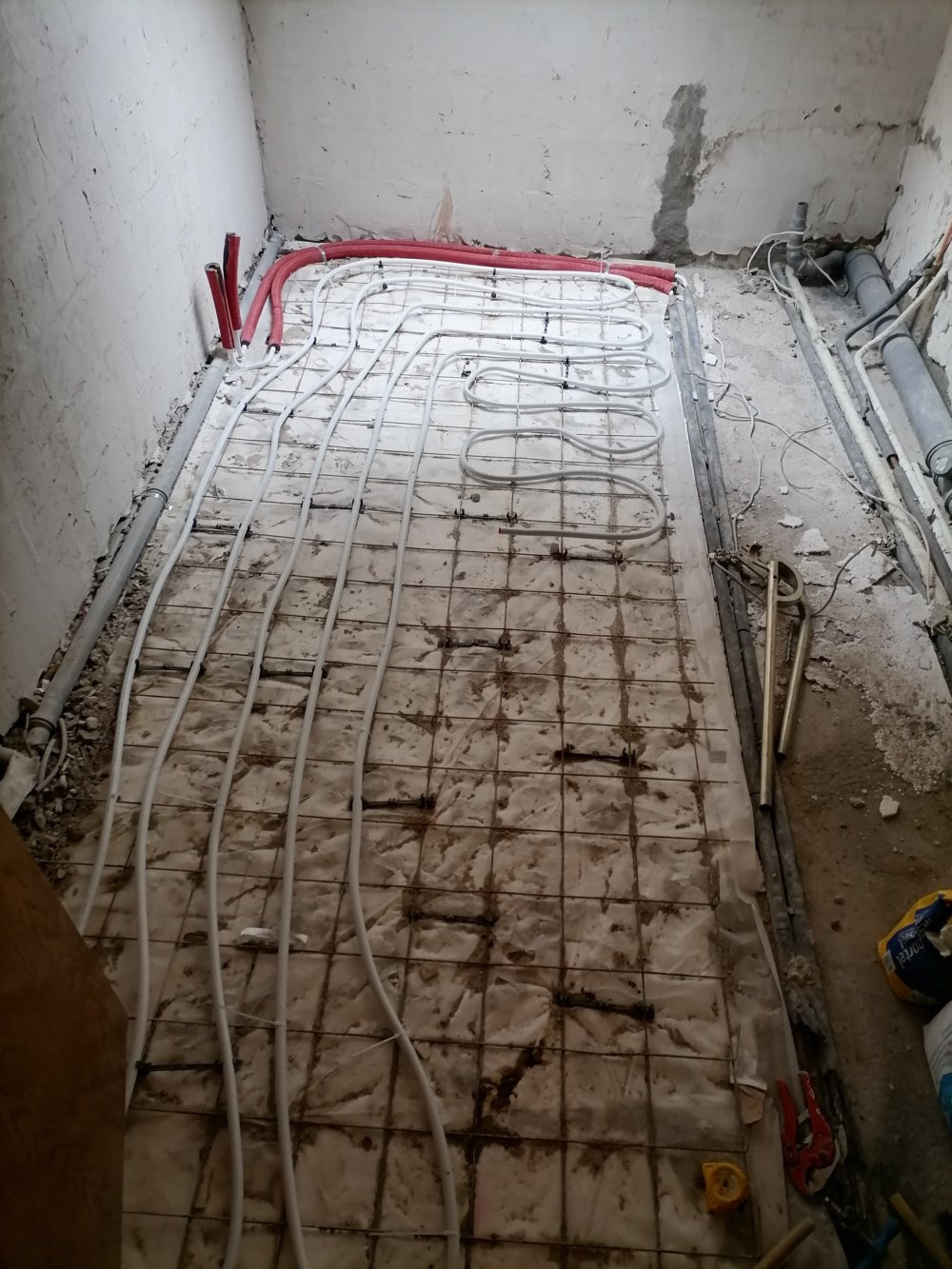 Montaż ogrzewania podłogowego: białe rurki układane na siatce zbrojeniowej w surowym pomieszczeniu, widoczne narzędzia hydrauliczne i fragment instalacji z czerwonymi rurami.