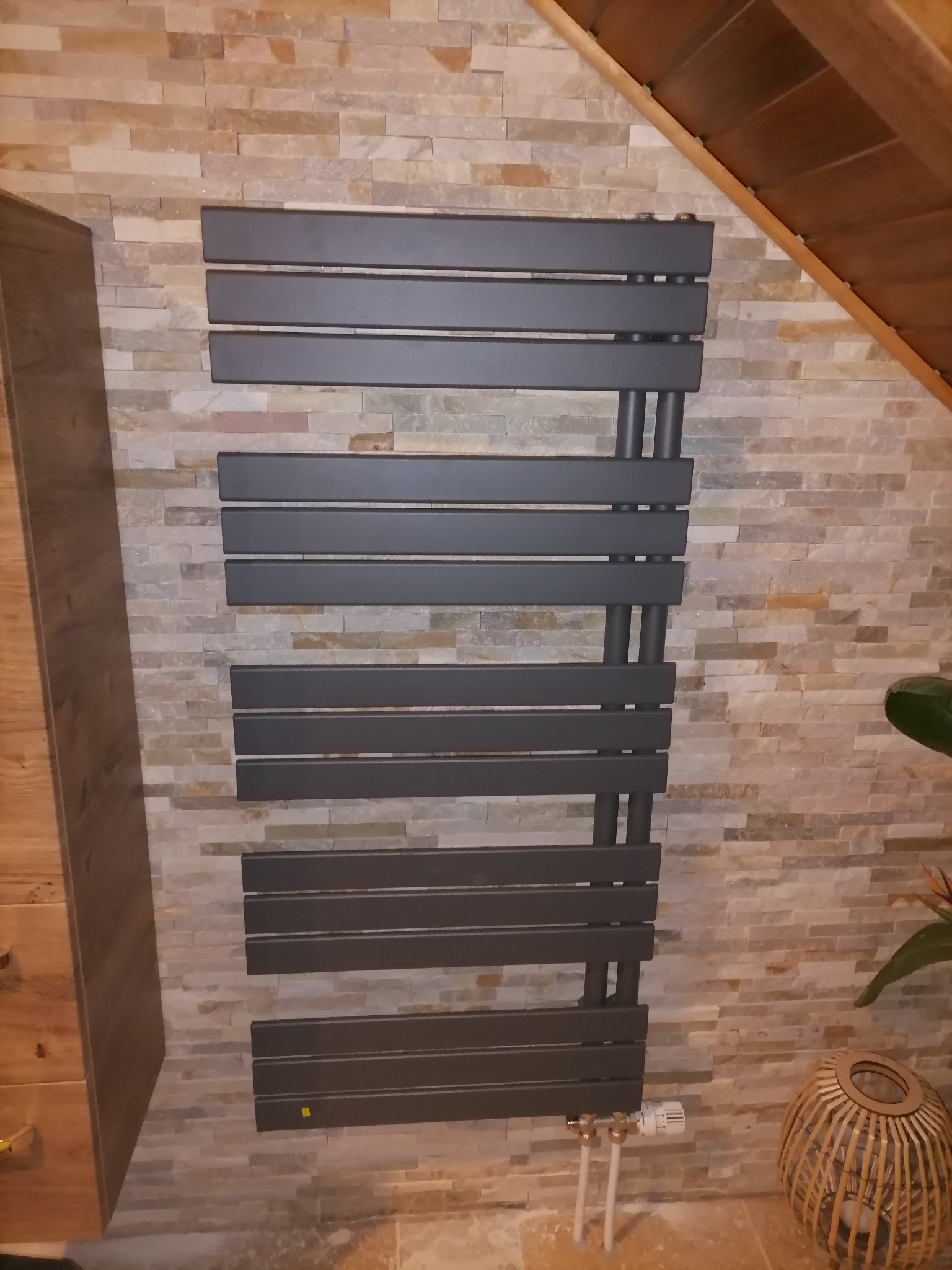 Szary, nowoczesny grzejnik drabinkowy zamontowany na ścianie z dekoracyjnej cegły, widoczne rury doprowadzające wodę i termostat, w tle drewniany strop i fragment szafki.