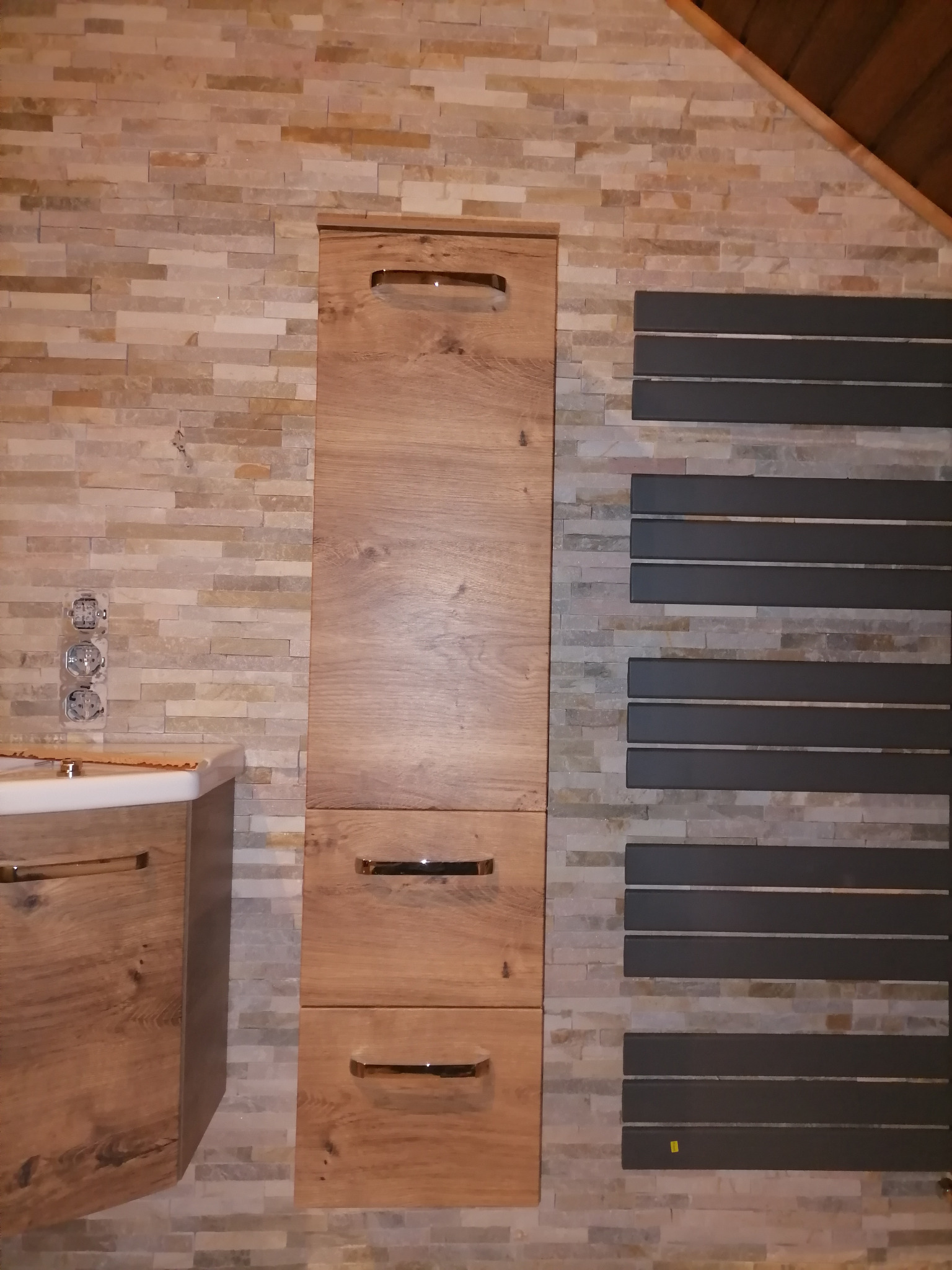 Nowoczesna łazienka z drewnianymi szafkami i grzejnikiem drabinkowym na ścianie z dekoracyjnej cegły, widoczne gniazdka elektryczne.