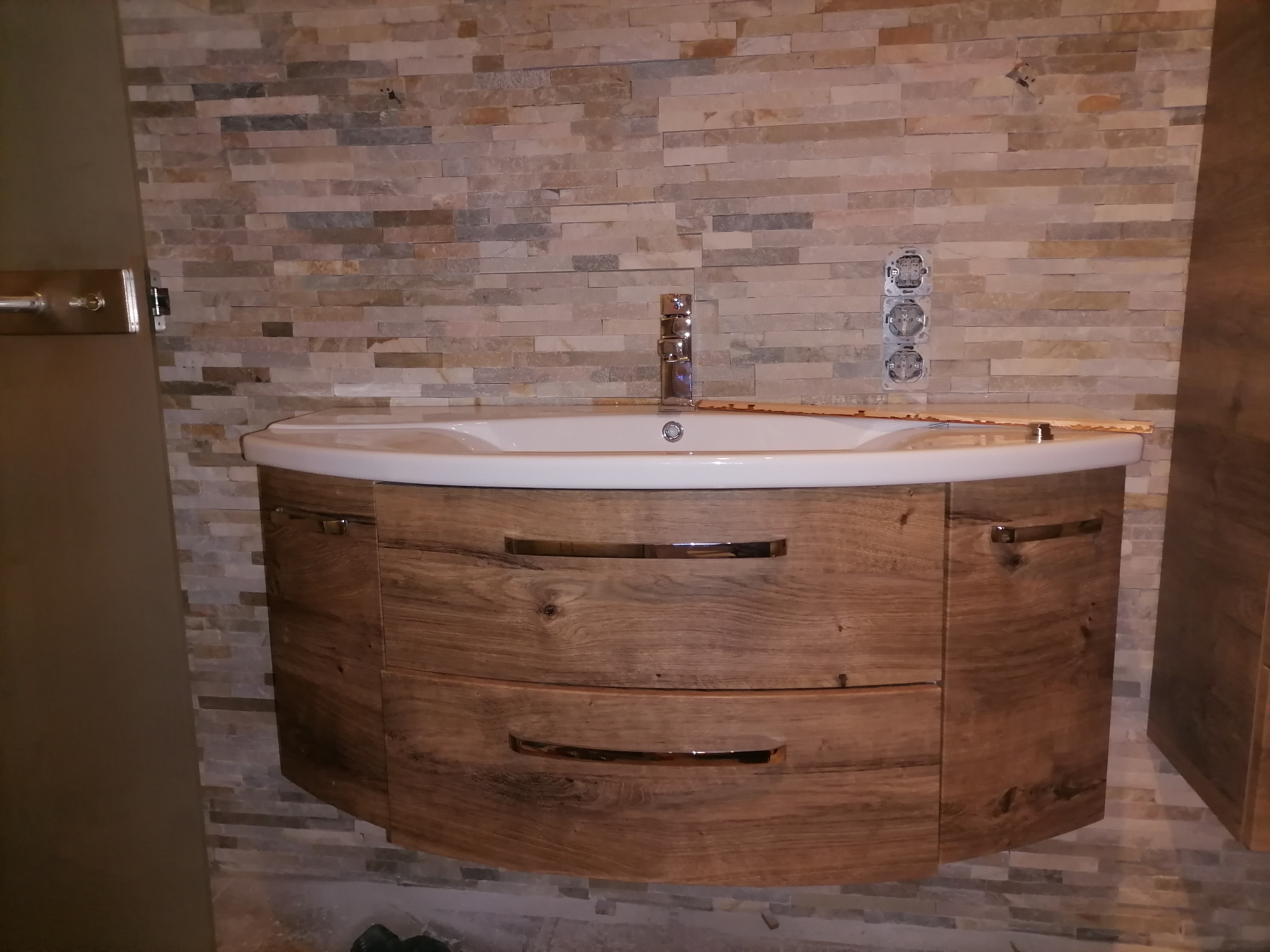 Nowoczesna umywalka z szafką z drewna w łazience z kamienną ścianą i widoczną instalacją elektryczną.