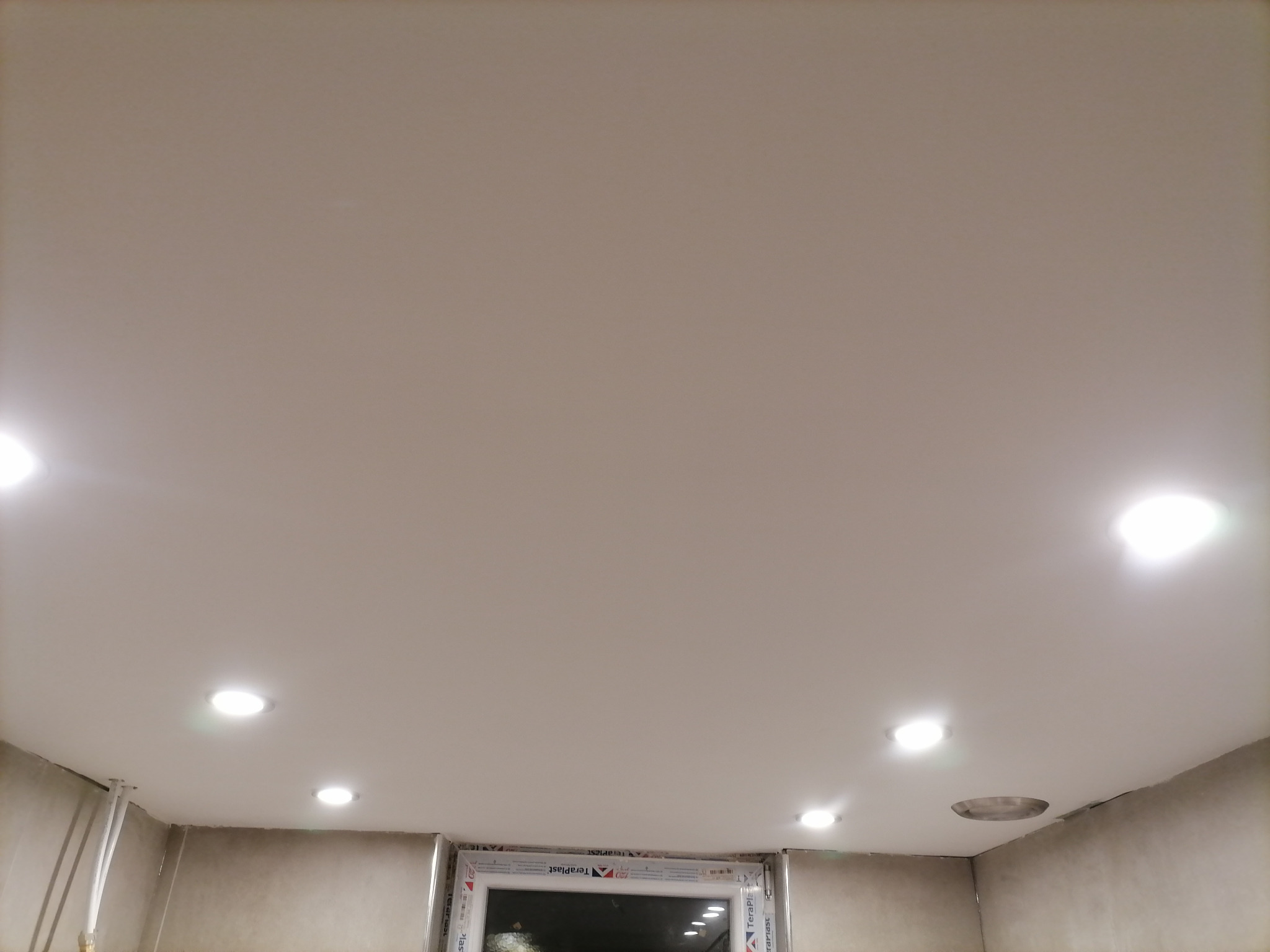 Biały sufit podwieszany z zamontowanymi punktowymi lampami LED, widoczne rury instalacji grzewczej przy ścianie oraz okno z białymi ramami.