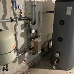 Zamontowany, szary bojler z podłączeniami hydraulicznymi, manometrami i zaworami. Widoczny zbiornik wyrównawczy IBO. Instalacja w surowym pomieszczeniu.