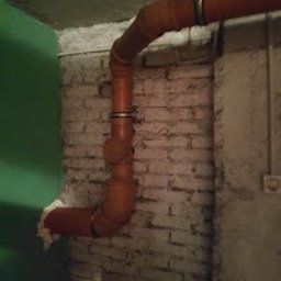 JTB SZYMON MICHALCZYK - Pomarańczowe rury kanalizacyjne z obejmami mocowane do starej, białej ściany z cegły w pomieszczeniu z zieloną ścianą i widoczną instalacją elektryczną.