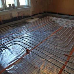 JTB SZYMON MICHALCZYK - Białe rury ogrzewania podłogowego ułożone na folii z siatką w pokoju, widoczne okno i fragment ściany. Instalacja przed wylaniem posadzki.