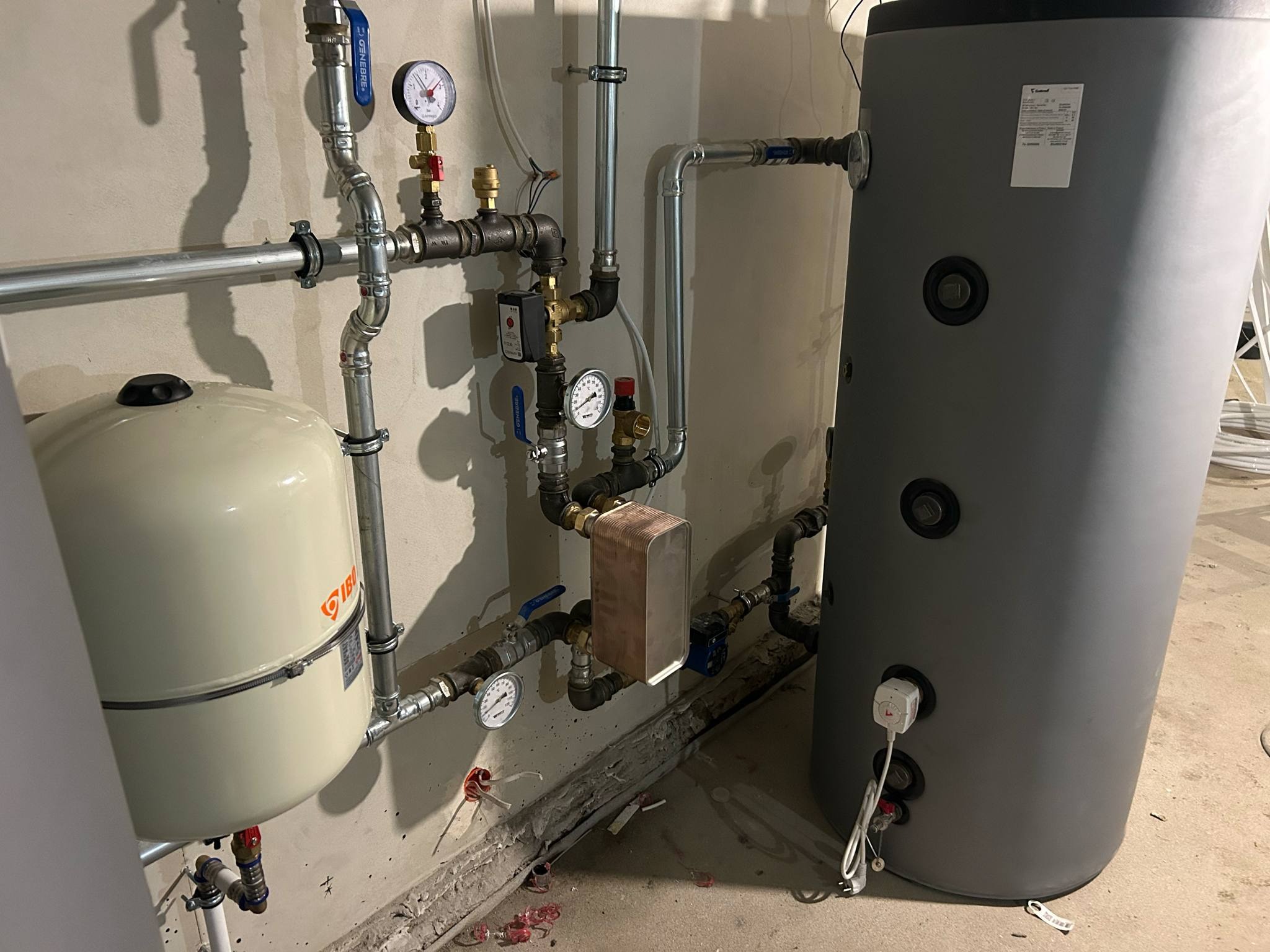 Zamontowany, szary bojler z podłączeniami hydraulicznymi, manometrami i zaworami. Widoczny zbiornik wyrównawczy IBO. Instalacja w surowym pomieszczeniu.