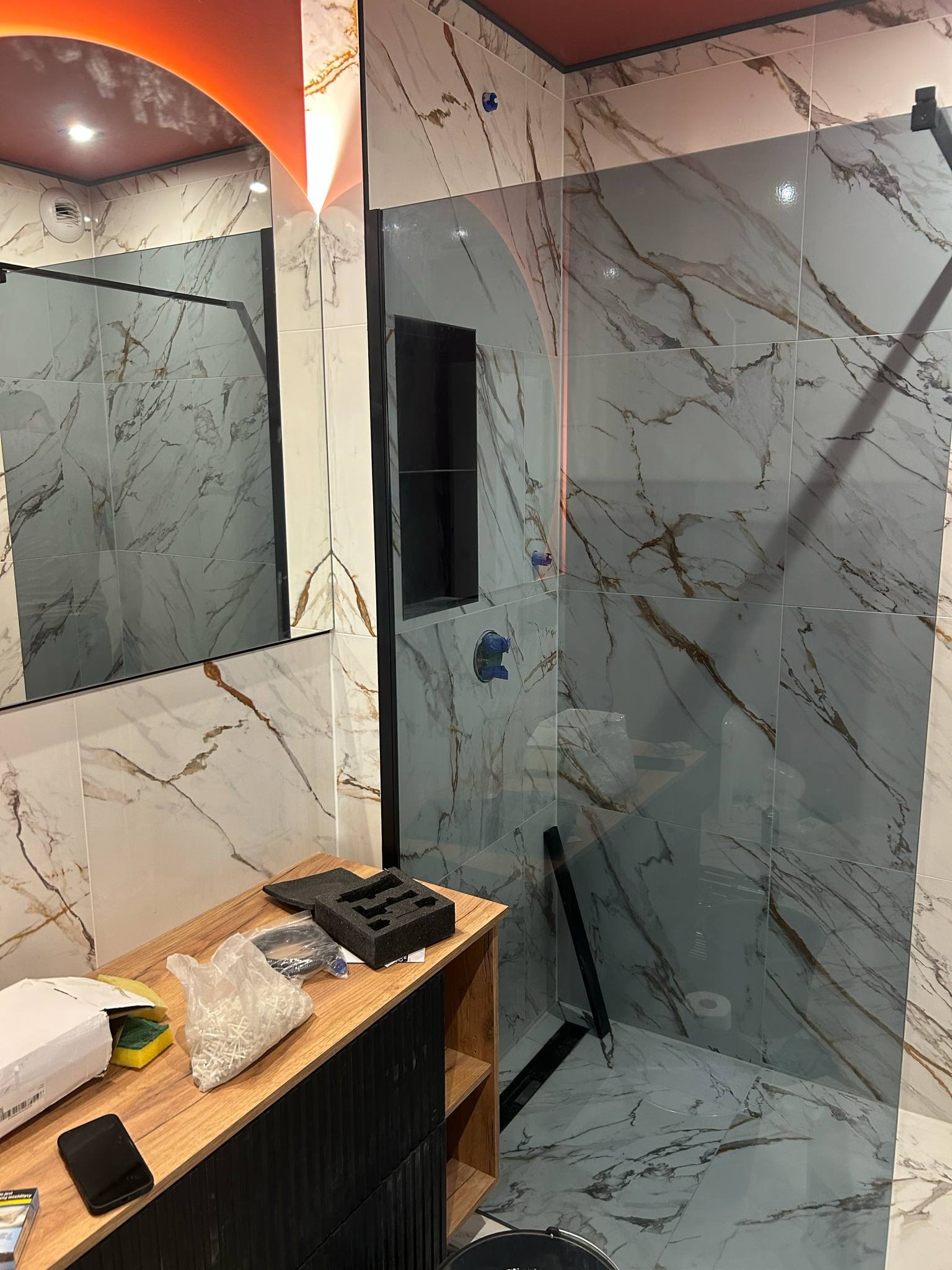 Nowoczesna kabina prysznicowa z marmurowymi płytkami i czarnymi akcentami. Widoczna półka i elementy montażowe. Oświetlenie sufitowe w ciepłej tonacji.