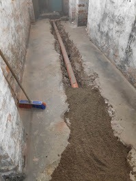 Rura kanalizacyjna w wykopie, zasypana piaskiem w surowym wnętrzu. Widoczna ściana z cegły i fragment miotły. Instalacja kanalizacyjna w trakcie prac.