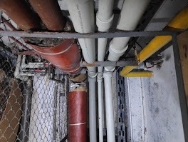 Różne rury (białe, brązowe, żółte) biegnące pod sufitem, wsparte na metalowej konstrukcji. Widoczne złączenia i izolacja. Część rur ukryta za siatką.