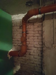 Pomarańczowe rury kanalizacyjne z obejmami mocowane do starej, białej ściany z cegły w pomieszczeniu z zieloną ścianą i widoczną instalacją elektryczną.