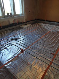 Białe rury ogrzewania podłogowego ułożone na folii z siatką w pokoju, widoczne okno i fragment ściany. Instalacja przed wylaniem posadzki.