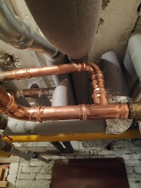 Miedziane rury instalacji centralnego ogrzewania pod sufitem, widoczne złącza i fragmenty izolacji, żółta rura w tle, surowy mur z cegły.