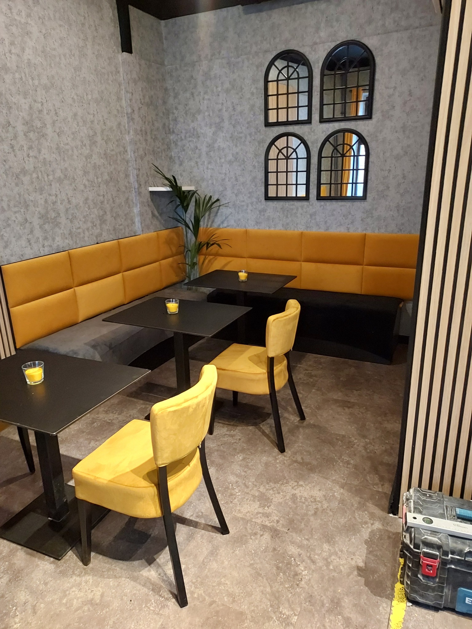 Wnętrze lokalu gastronomicznego z żółtymi krzesłami i kanapą, czarnymi stolikami z dekoracyjnymi świeczkami, szarą ścianą z lustrami w czarnych ramach oraz skrzynką narzędziową w rogu.