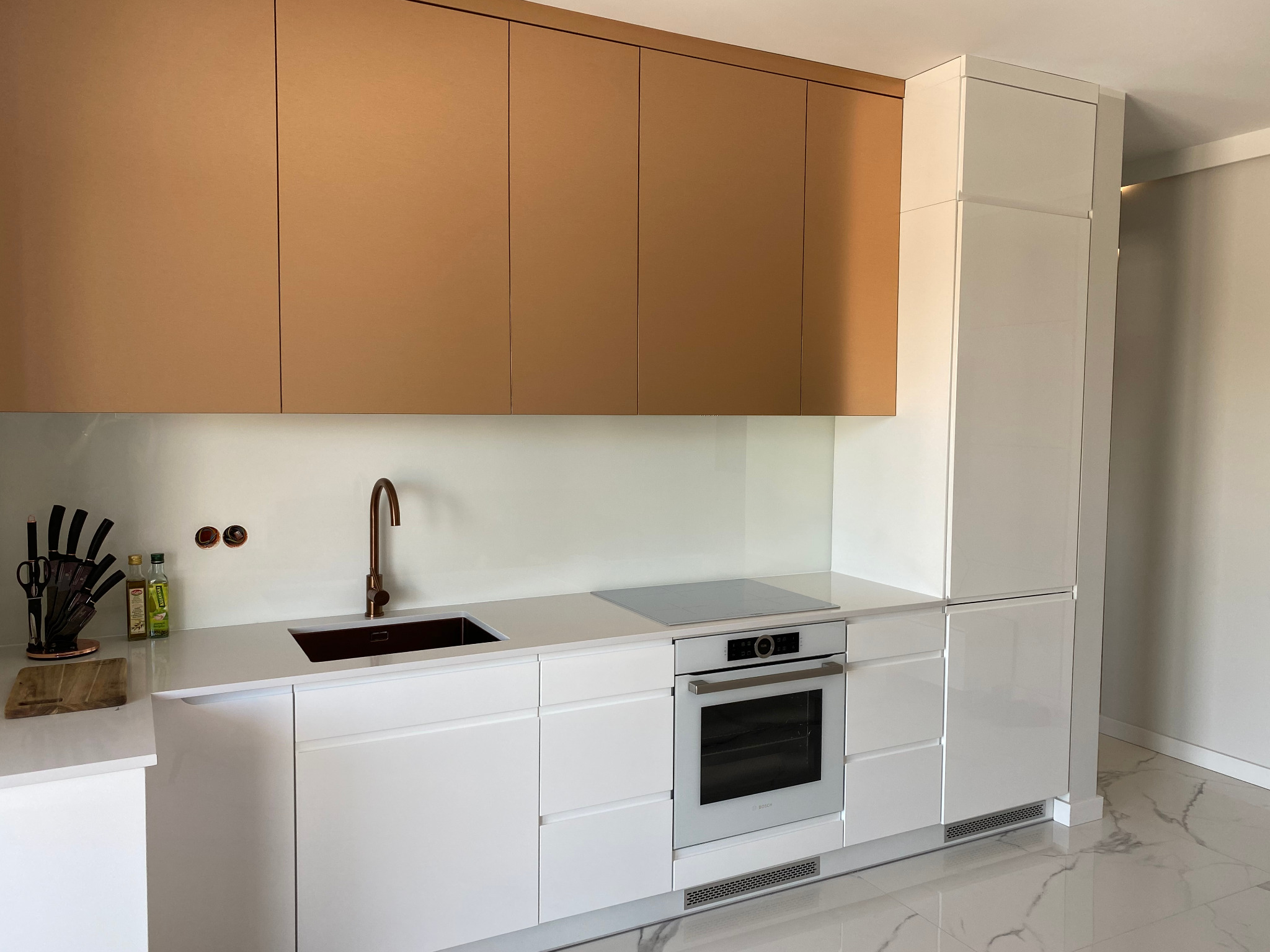 Nowoczesna kuchnia z białymi szafkami dolnymi i górnymi w odcieniu miedzianym, miedziana bateria i zlew, biały blat, płyta indukcyjna i piekarnik Bosch, na podłodze biały marmur.