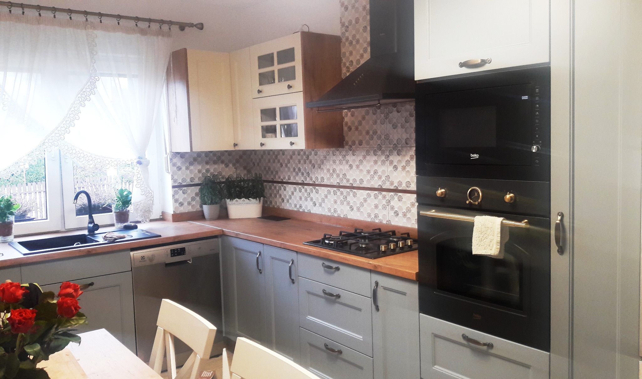 Szara kuchnia z drewnianym blatem, czarnym piekarnikiem i płytą gazową, widok na okno z dekoracyjną firanką i zlewozmywak, kwiaty w wazonie na stole