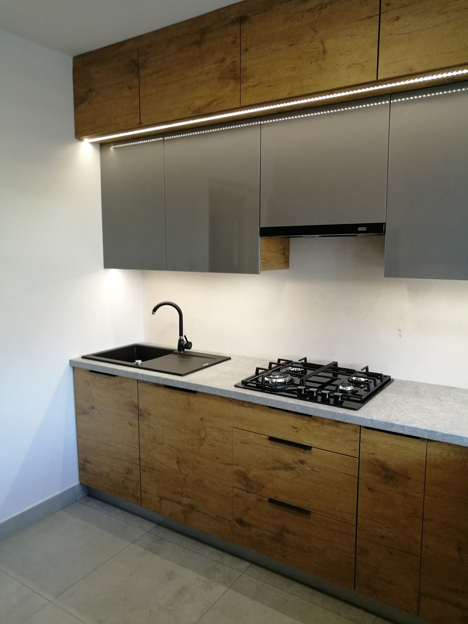 Nowoczesna kuchnia z szarymi szafkami górnymi i drewnopodobnymi szafkami dolnymi, czarnym zlewem i płytą gazową, oświetleniem LED pod szafkami górnymi.