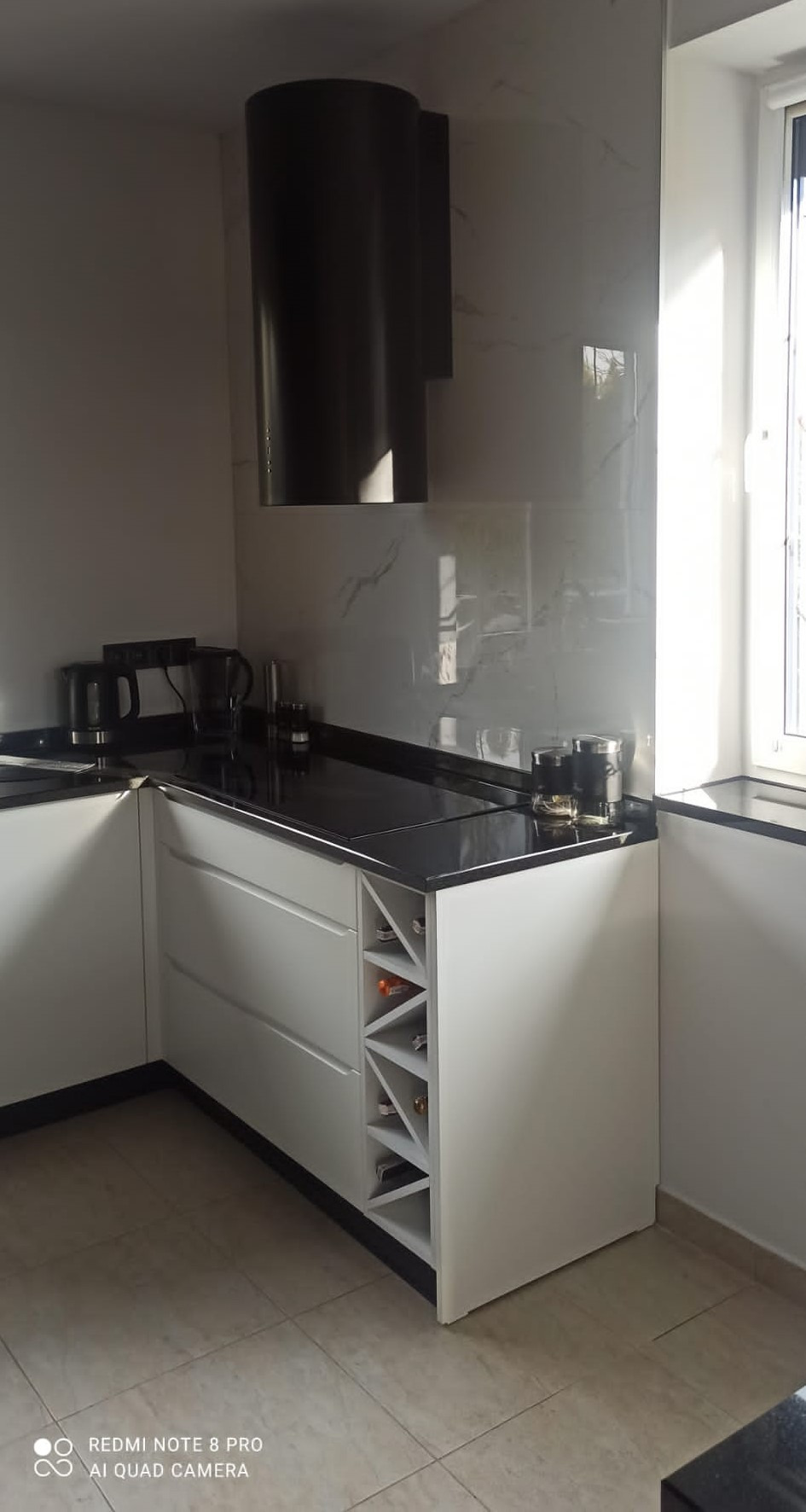 Nowoczesna kuchnia z białymi szafkami, czarnym blatem i cylindrycznym okapem, uzupełniona o wbudowany regał na wino.