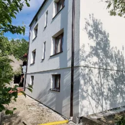 Dwupiętrowy budynek z jasną elewacją, brązowymi oknami i rynnami, z widocznym cieniem drzewa na ścianie oraz żółtym wężem na ziemi.