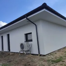 Świeżo otynkowany, biały budynek z czarną stolarką okienną i orynnowaniem, klimatyzator Kaisai na zewnątrz, widoczny fragment niezagospodarowanego terenu.