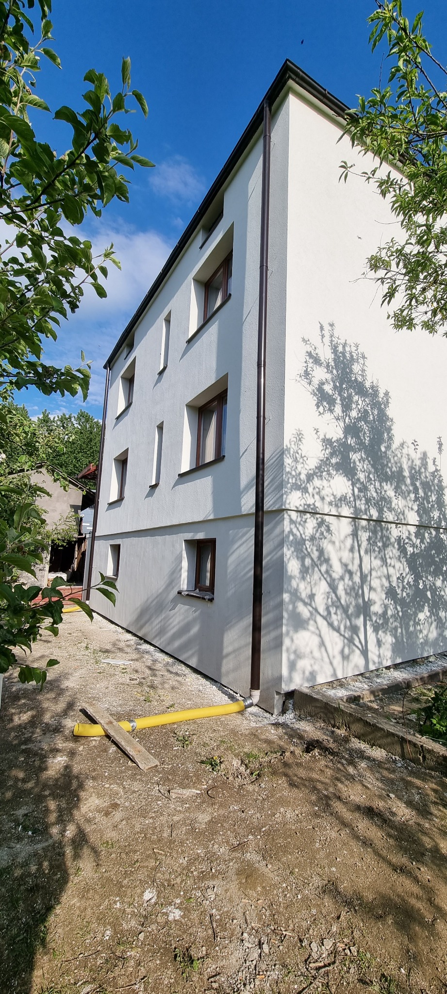 Dwupiętrowy budynek z jasną elewacją, brązowymi oknami i rynnami, z widocznym cieniem drzewa na ścianie oraz żółtym wężem na ziemi.