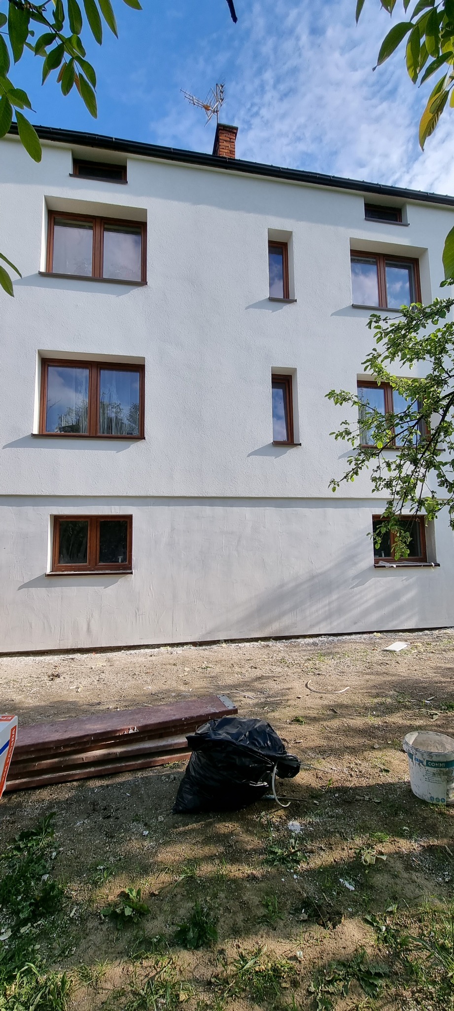 Odnowiona biała elewacja budynku z brązowymi oknami, widoczne ślady prac wykończeniowych, w tle błękitne niebo i zielone liście drzew.