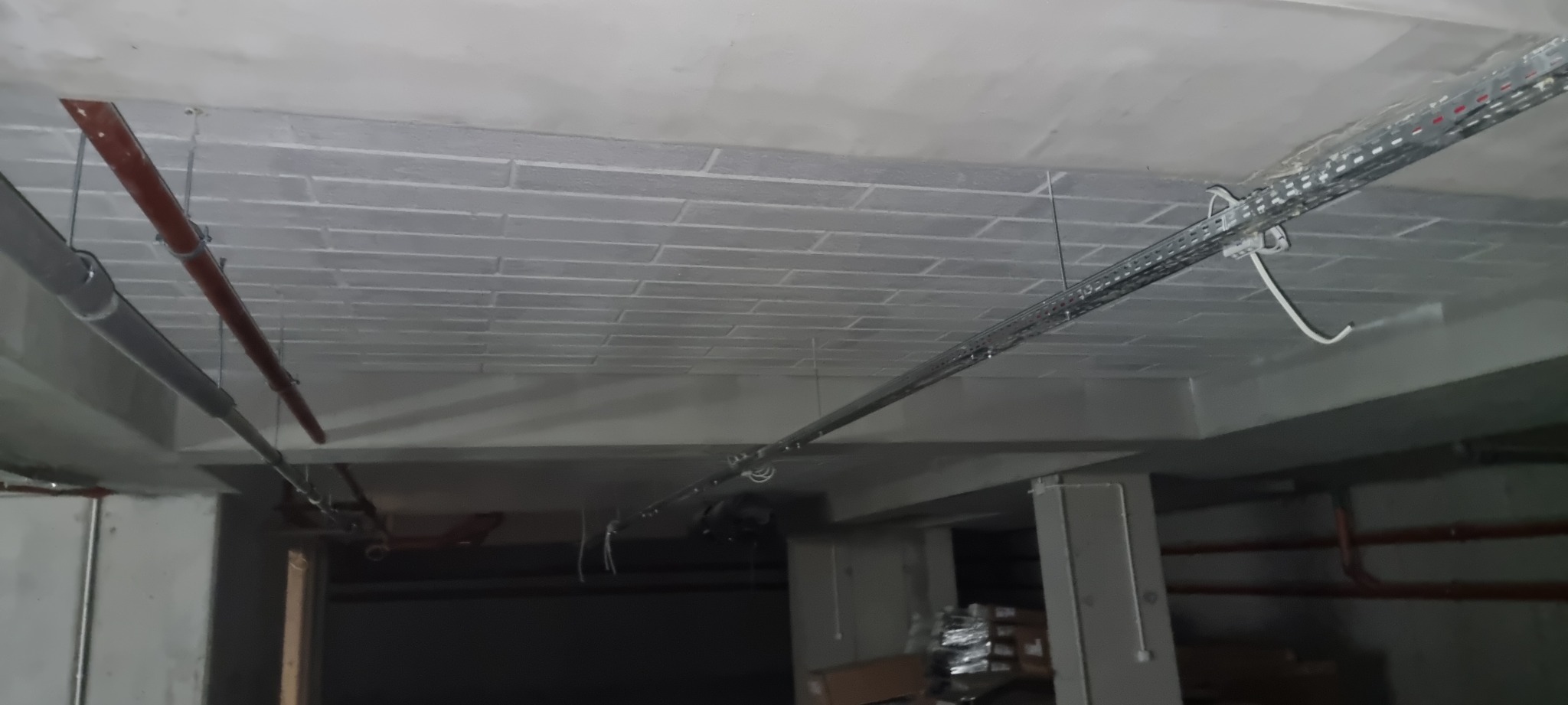 Szare, pomalowane cegły na suficie piwnicy lub parkingu podziemnego z widocznymi rurami i korytami kablowymi.