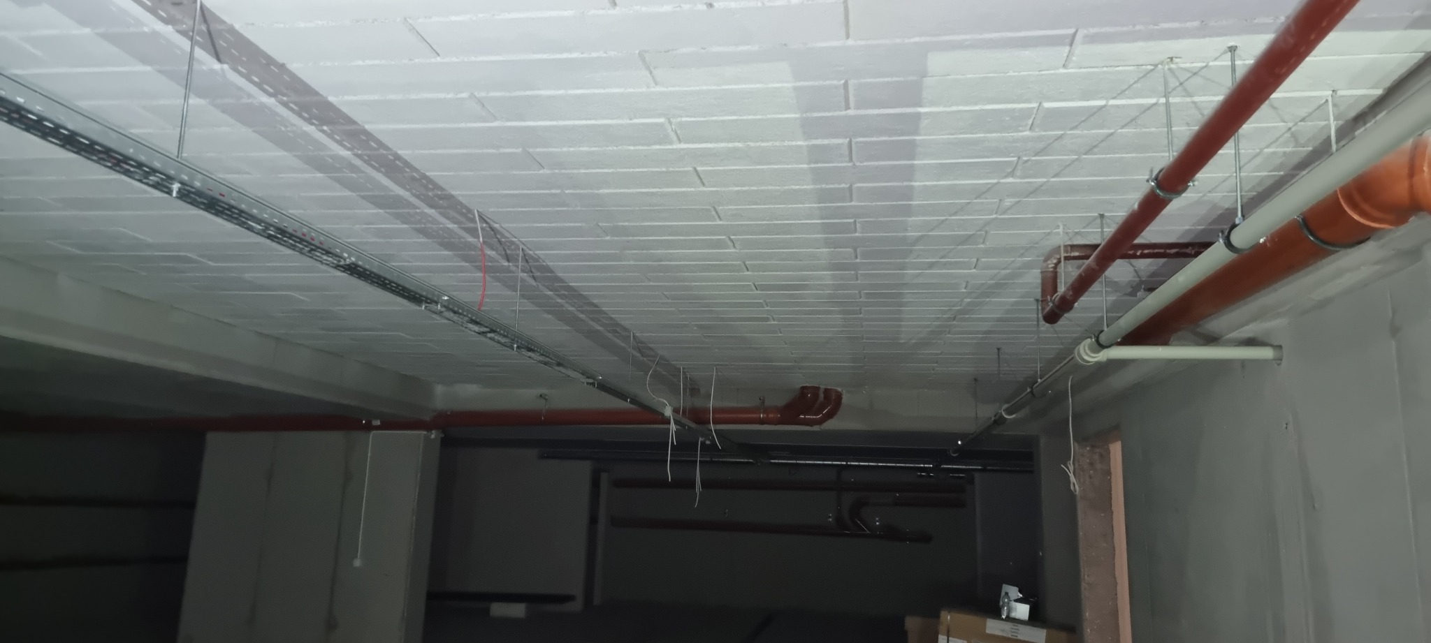 Szare, betonowe ściany i sufit z widocznymi instalacjami: rurami w kolorze ceglastym, metalowymi korytkami kablowymi i zwisającymi przewodami elektrycznymi.