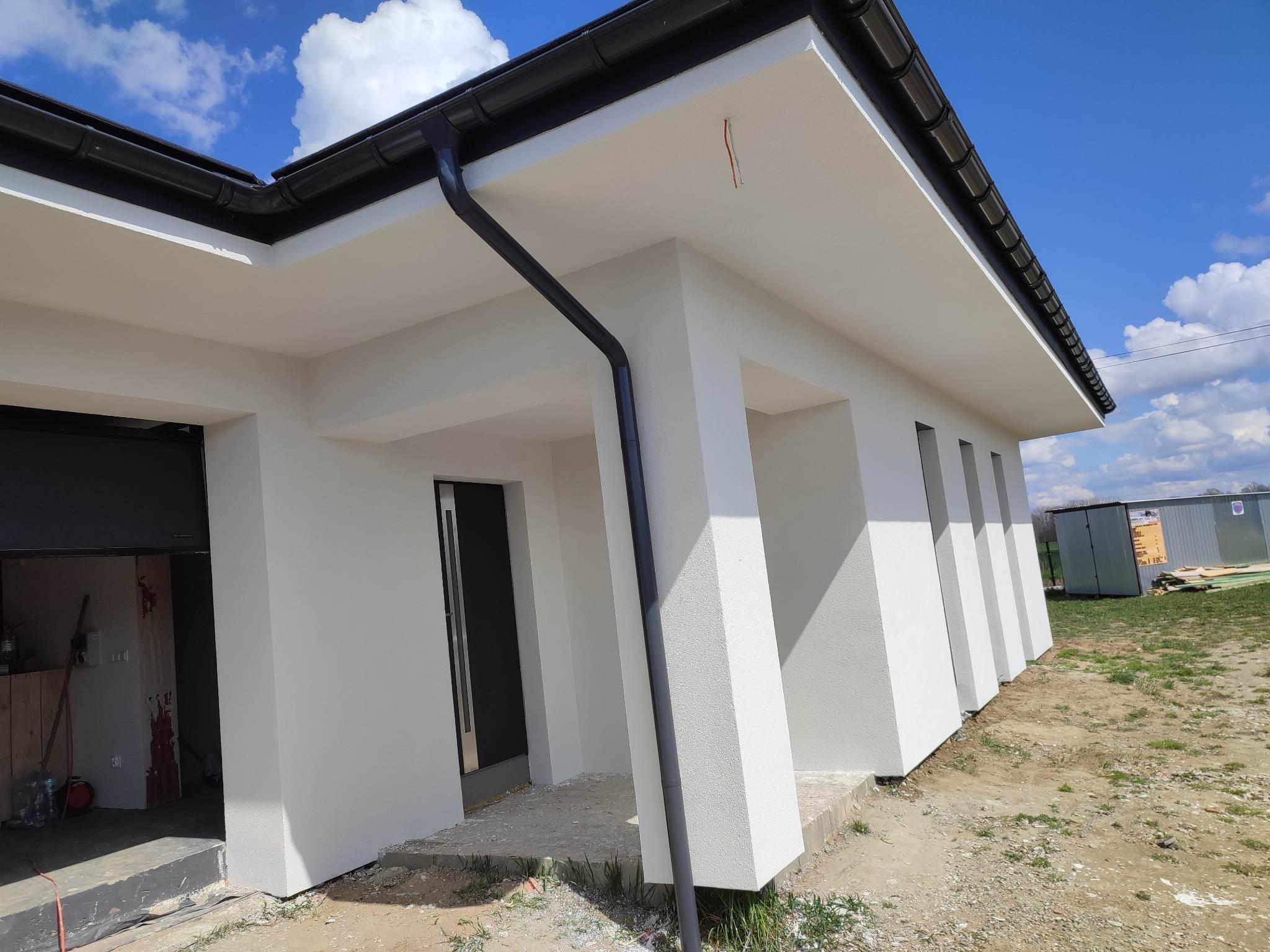 Nowoczesny dom jednorodzinny z białą elewacją, czarną stolarką okienną i orynnowaniem, widoczne niedokończone wykończenia terenu wokół budynku oraz otwarte pomieszczenie garażowe z narzędziami.