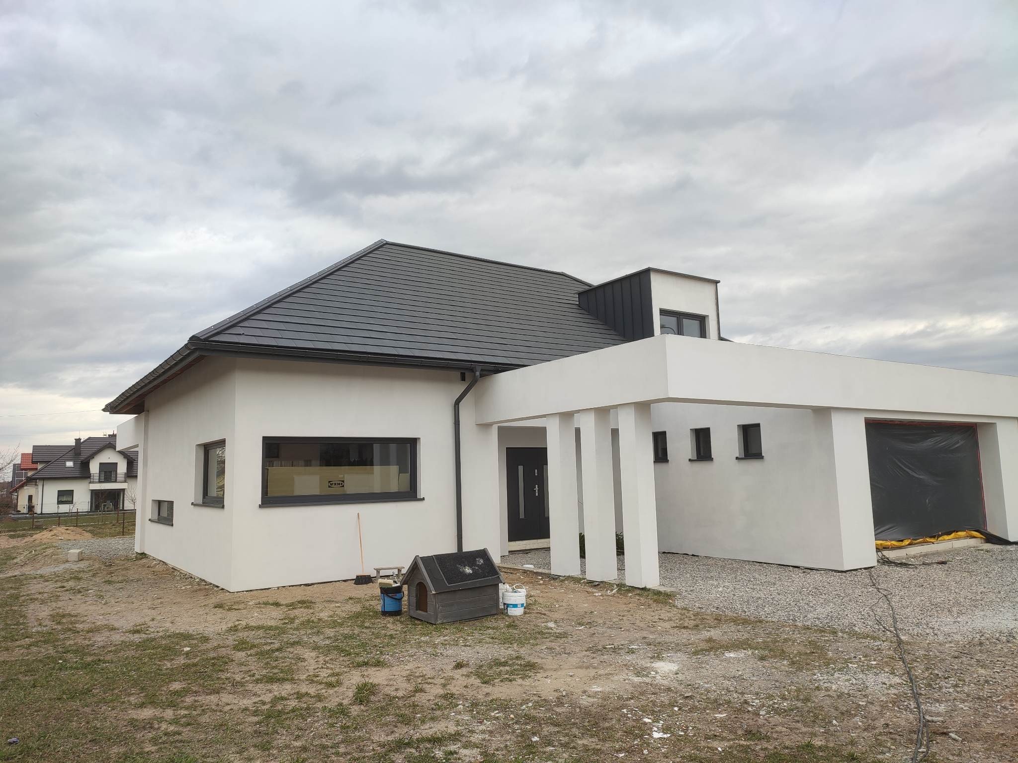 Nowoczesny dom jednorodzinny z płaskim dachem i białymi ścianami, garaż w trakcie wykończenia, widoczne akcesoria malarskie i buda dla psa na posesji.