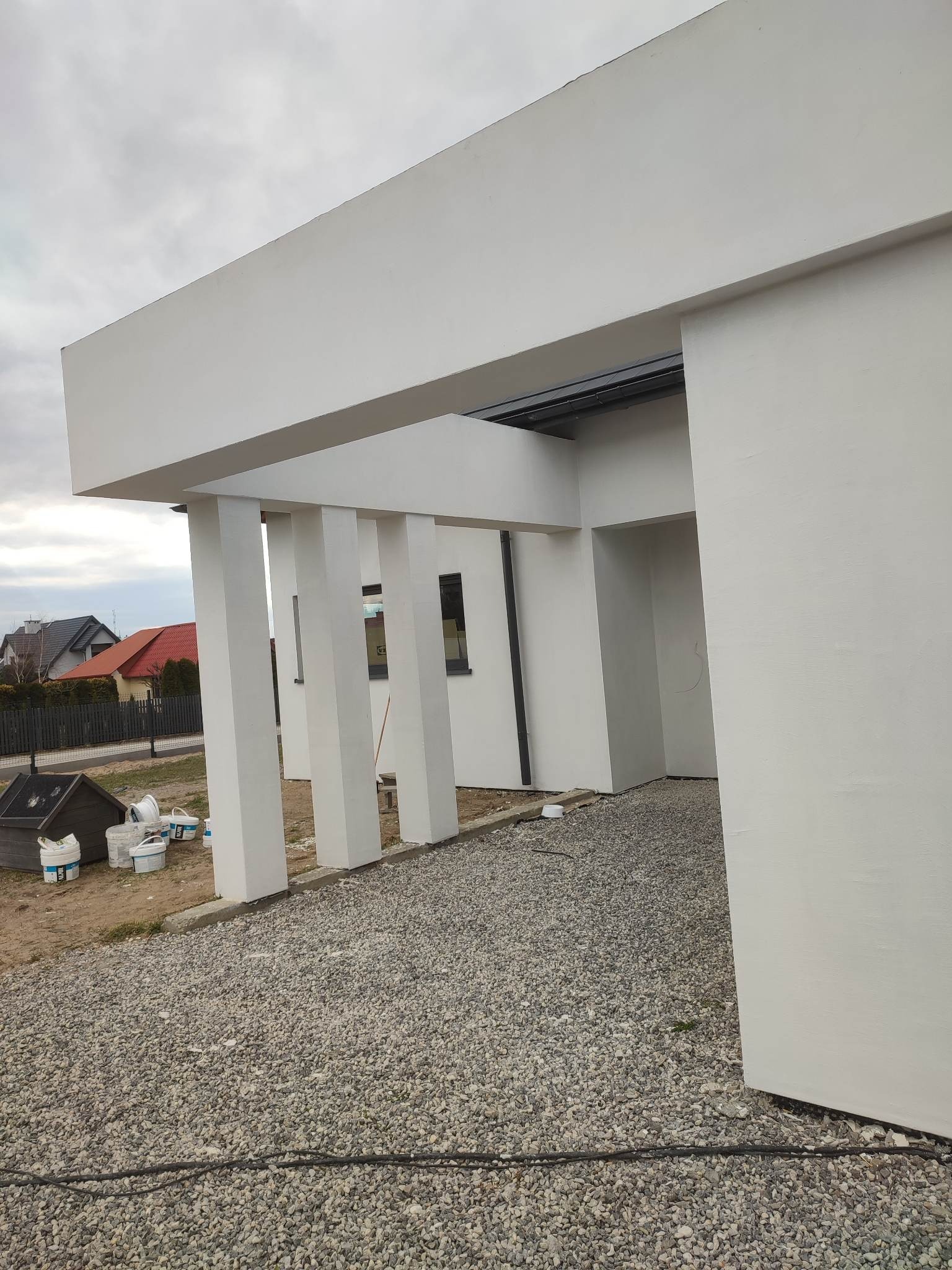 Nowoczesny dom jednorodzinny z białymi ścianami i kolumnami, świeżo po tynkowaniu, widoczne wiaderka po farbie i żwir na podłożu.